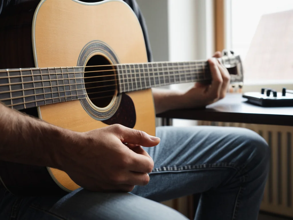 A beginner guitarist sitting at a desk with an acoustic guitar practicing chords and scales.